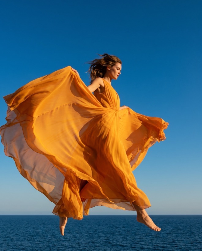 A dancer in a saffron gown leaping against a clear blue sky above the sea